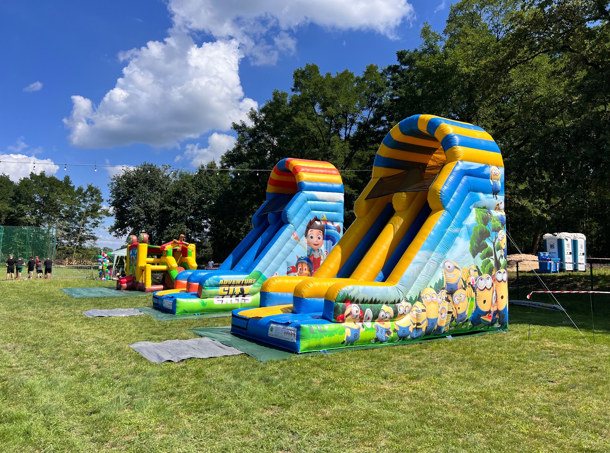 Dwa dmuchane zamki ze zjeżdżalniami, jeden z motywem Minionków, drugi z postaciami z bajki Psi Patrol, rozstawione na trawie podczas słonecznego dnia.