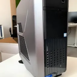 Stacjonarny komputer marki Alienware w srebrno-czarnej obudowie, stojący na biurku w jasnym pomieszczeniu biurowym. Widoczny z boku i przodu, z logo na obudowie.