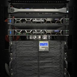 MKS Polska Rusinek Spółka Jawna - Rząd serwerów Dell EMC w szafie rack, widoczne diody i wyświetlacze. Na dole UPS z wyświetlaczem napięcia 231V. Kable zasilające z tyłu. Oświetlenie LED w szafie.