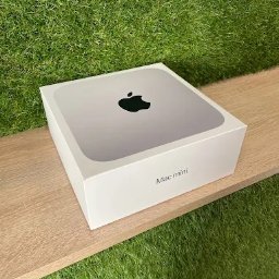MKS Polska Rusinek Spółka Jawna - Pudełko komputera Mac mini na drewnianej półce, z zielonym trawiastym tłem. Widoczne logo Apple i napis 'Mac mini'.