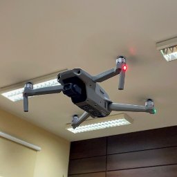 MKS Polska Rusinek Spółka Jawna - Szary dron Mavic Air 2 w biurze, blisko sufitu z lampami. Widoczne czerwone i zielone światła sygnalizacyjne na ramionach drona. Zastosowanie w monitoringu lub inspekcji.