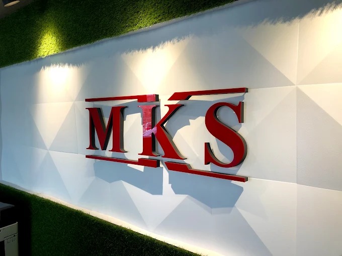 Logo 'MKS' w kolorze czerwonym, umieszczone na białej ścianie z geometrycznym wzorem, oświetlone punktowo od góry. Elementy dekoracyjne z zielonej trawy.