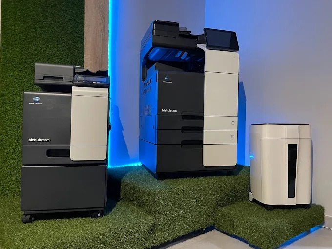 Dwa nowoczesne kserokopiarki Konica Minolta Bizhub w biurze, z podświetleniem LED i niszczarką dokumentów na zielonym podwyższeniu.