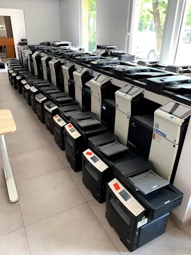 Rząd używanych kserokopiarek A4 i A3 różnych modeli, ustawionych w rzędach w jasnym pomieszczeniu z oknami. Widoczne różne marki i konfiguracje urządzeń.