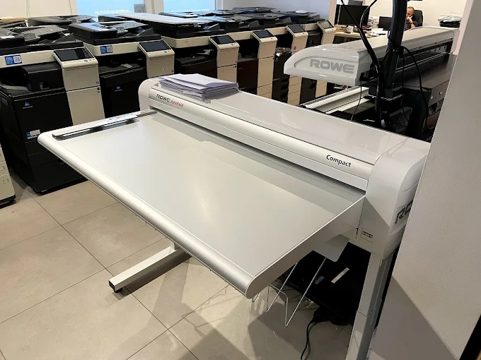 Szerokoformatowy skaner ROWE Compact z podajnikiem na dokumenty, widoczny w otoczeniu innych urządzeń biurowych, na jasnej podłodze. Ujęcie z bliska, z boku.