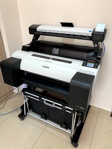 Duża, biało-czarna kserokopiarka Canon A0 na kółkach, z widocznym panelem sterowania i podajnikiem papieru, ustawiona w jasnym pomieszczeniu biurowym.