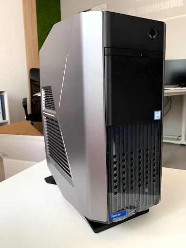 Stacjonarny komputer marki Alienware w srebrno-czarnej obudowie, stojący na biurku w jasnym pomieszczeniu biurowym. Widoczny z boku i przodu, z logo na obudowie.