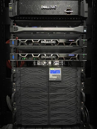 Rząd serwerów Dell EMC w szafie rack, widoczne diody i wyświetlacze. Na dole UPS z wyświetlaczem napięcia 231V. Kable zasilające z tyłu. Oświetlenie LED w szafie.