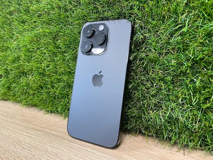 Grafitowy iPhone Pro z trzema obiektywami aparatu, leżący na sztucznej trawie i drewnianej powierzchni. Widoczny tył telefonu z logo Apple.