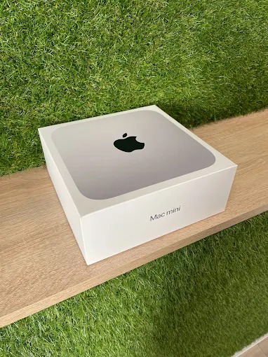 Pudełko komputera Mac mini na drewnianej półce, z zielonym trawiastym tłem. Widoczne logo Apple i napis 'Mac mini'.