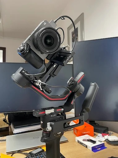Aparat Sony Alpha z obiektywem zamocowany na stabilizatorze DJI Ronin RS 3, ustawiony na biurku z monitorem i akcesoriami. Sprzęt foto-wideo w gotowości.