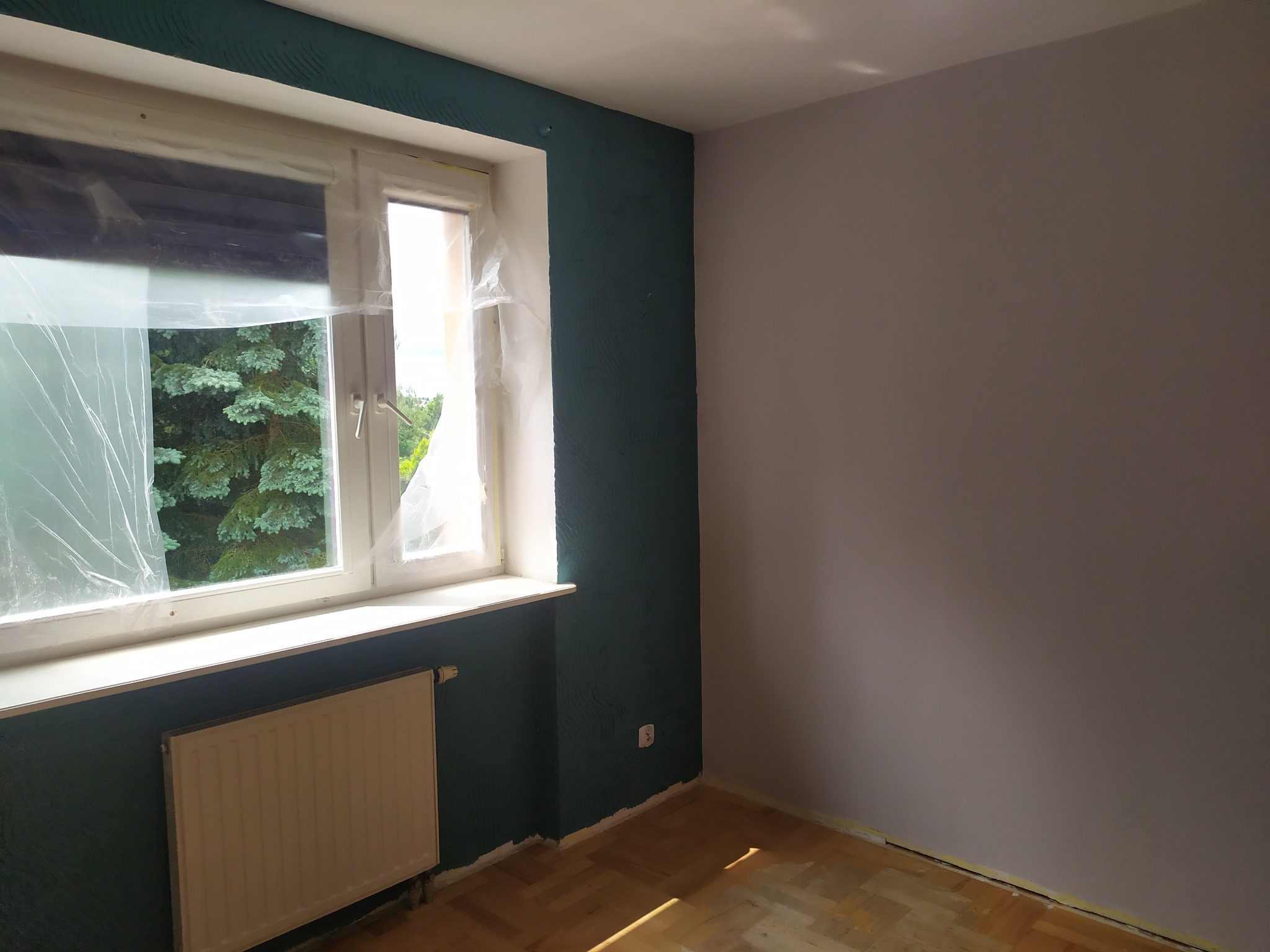 Pokój po malowaniu: ściana w odcieniu zieleni i szarości, okno zabezpieczone folią. Widoczny grzejnik i fragment drewnianej podłogi. Jasne wnętrze.
