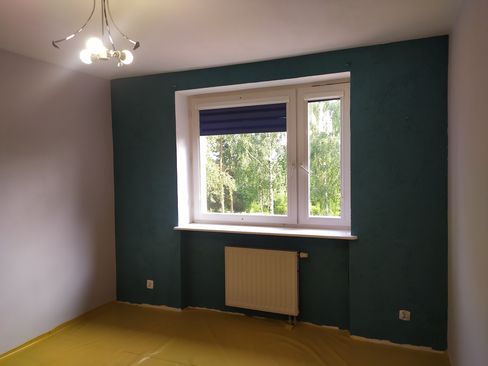 Pokój po malowaniu: ściany w odcieniach zieleni, okno z roletą, grzejnik. Podłoga zabezpieczona folią malarską. Widok na drzewa za oknem.
