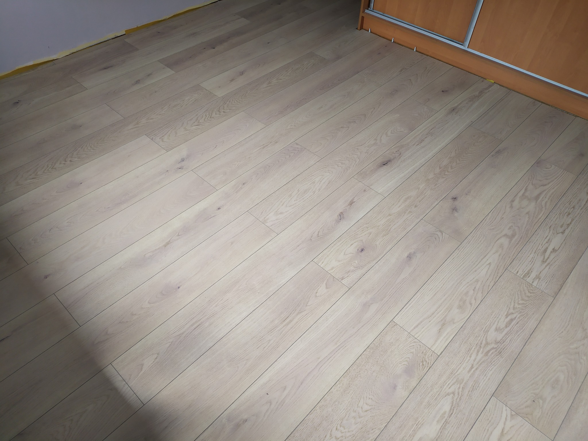 Jasne panele podłogowe ułożone wzdłuż ściany z zabudową, widoczne taśmy zabezpieczające przy ścianie, ukazujące świeżo ukończony montaż podłogi.