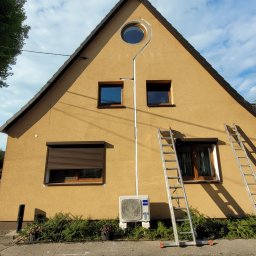 MEGAZORD - Żółty dom z zamontowanym klimatyzatorem Haier na elewacji. Dwie drabiny opierają się o ścianę. Instalacja w korytkach. Widok z dołu, błękitne niebo.