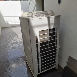 MEGAZORD - Biała jednostka zewnętrzna klimatyzacji Daikin VRV III na zewnątrz budynku, z widoczną kratką wentylacyjną i elementami konstrukcyjnymi. Tło stanowi jasna ściana i ciemna podłoga.