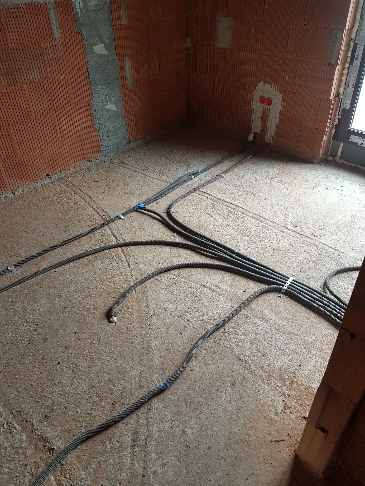 Wnętrze w trakcie instalacji elektrycznej: kable w peszlach prowadzone po podłodze w surowym stanie, ściany z czerwonej cegły dziurawki, widoczne puszki elektryczne.