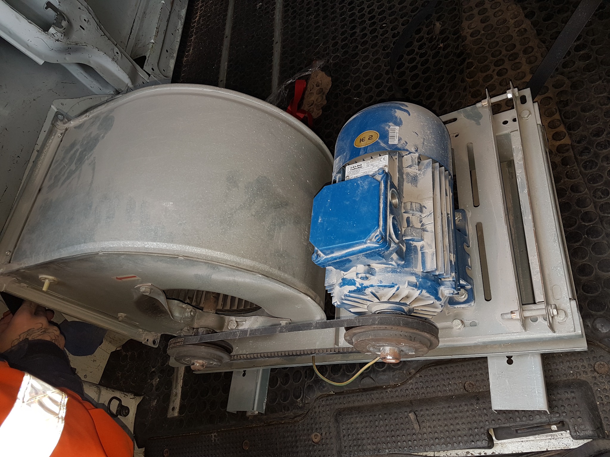 Wentylator przemysłowy z silnikiem elektrycznym, połączone paskiem klinowym, zamontowane na metalowej konstrukcji wewnątrz pojazdu serwisowego. Widoczne ślady użytkowania.