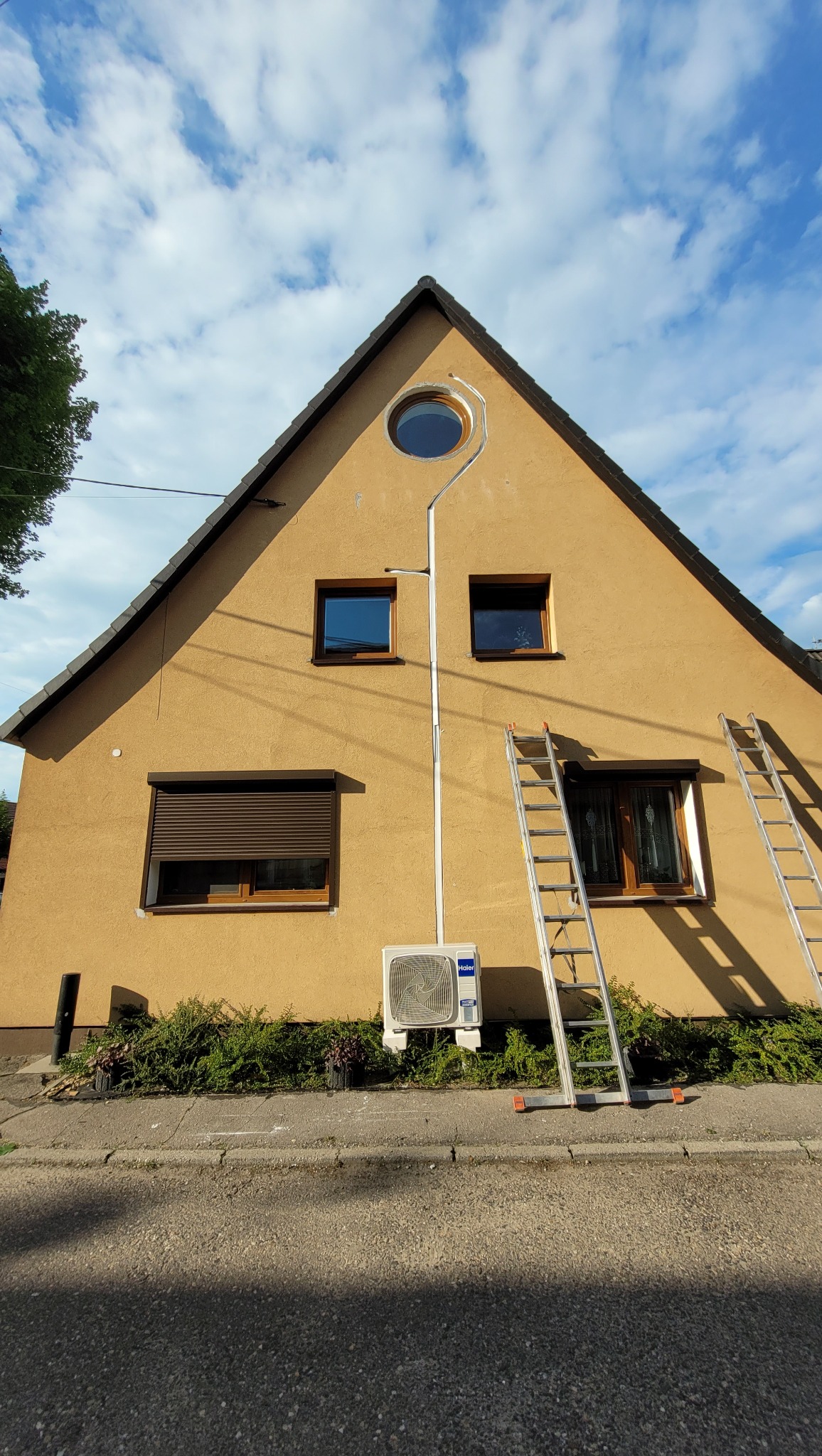 Żółty dom z zamontowanym klimatyzatorem Haier na elewacji. Dwie drabiny opierają się o ścianę. Instalacja w korytkach. Widok z dołu, błękitne niebo.
