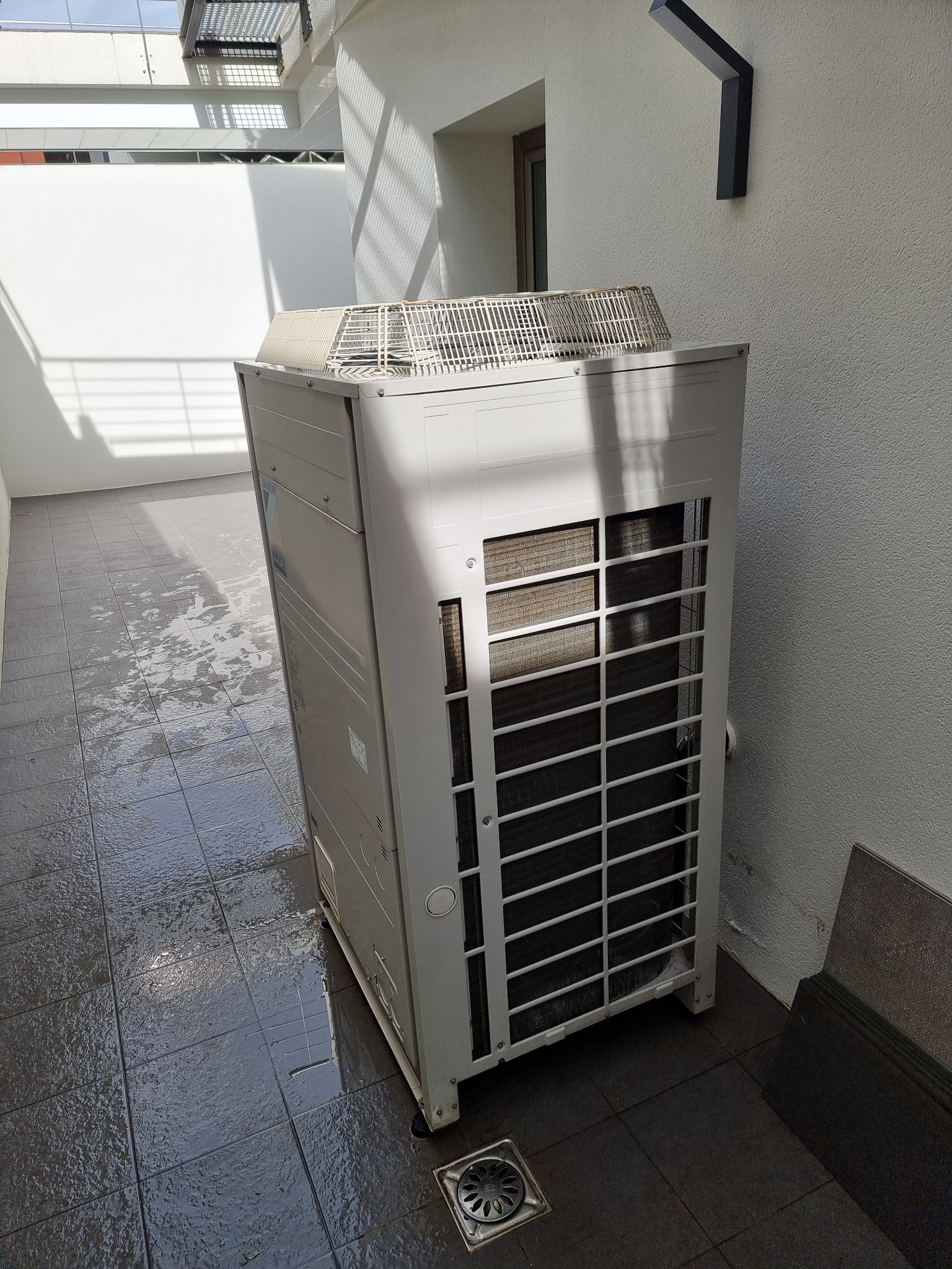 Biała jednostka zewnętrzna klimatyzacji Daikin VRV III na zewnątrz budynku, z widoczną kratką wentylacyjną i elementami konstrukcyjnymi. Tło stanowi jasna ściana i ciemna podłoga.