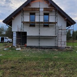 Wykończenia wnętrz - Dom jednorodzinny w trakcie prac wykończeniowych elewacji, z widocznym rusztowaniem. Ściany pokryte świeżym tynkiem, okna w drewnianej ramie.