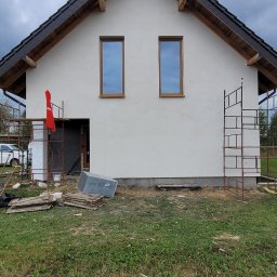 Wykończenia wnętrz - Dom jednorodzinny w trakcie prac wykończeniowych. Widoczne rusztowania, świeżo otynkowana elewacja i materiały budowlane na trawniku.
