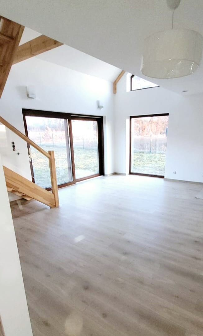 Jasne wnętrze domu z drewnianymi schodami i dużymi oknami przesuwnymi. Parkiet w odcieniach szarości, białe ściany i sufit. Widoczne drewniane elementy konstrukcyjne.