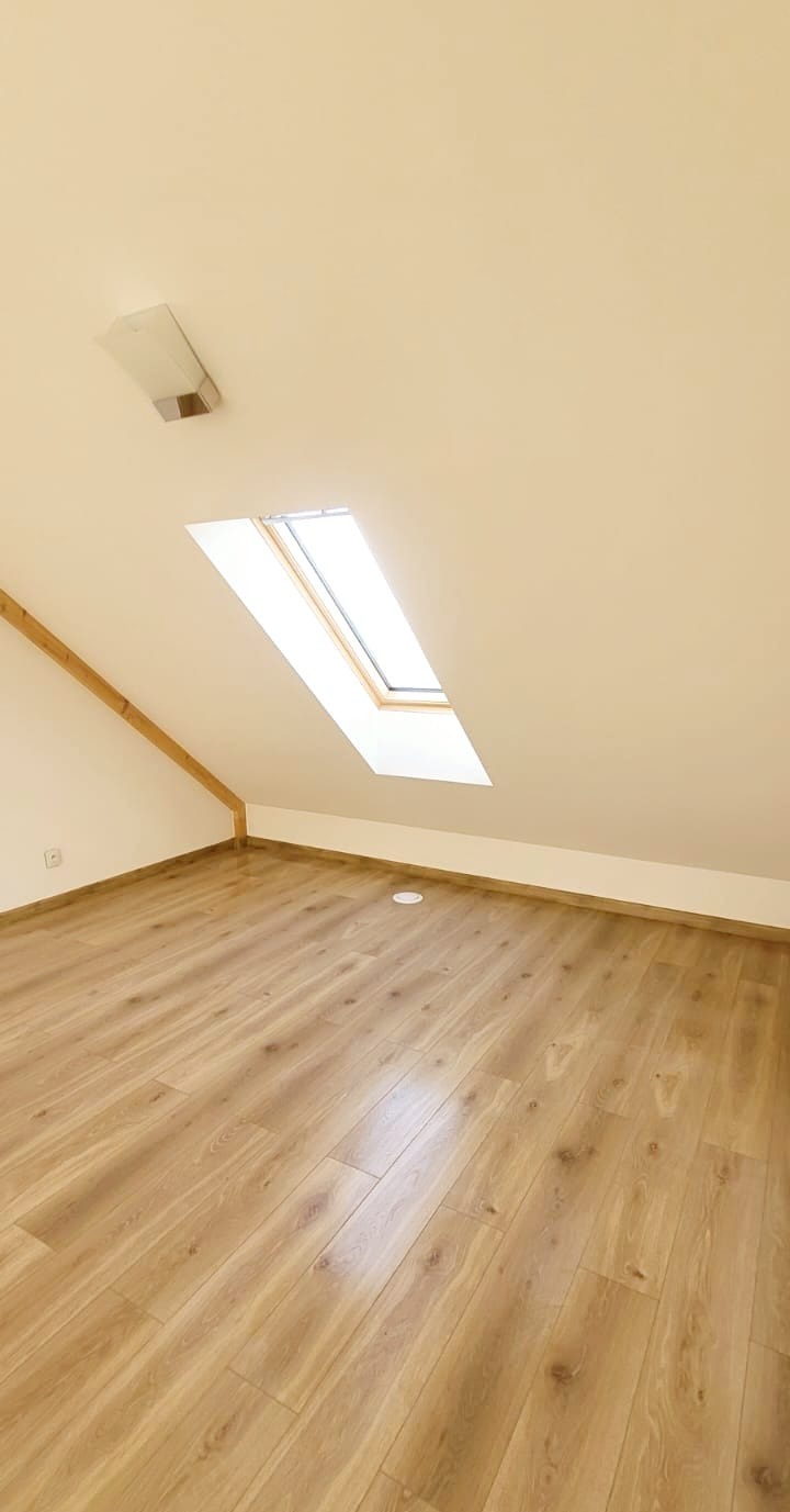 Jasne panele podłogowe w nowym pokoju na poddaszu z oknem dachowym i skosem. Widoczna listwa przypodłogowa i lampa na suficie.
