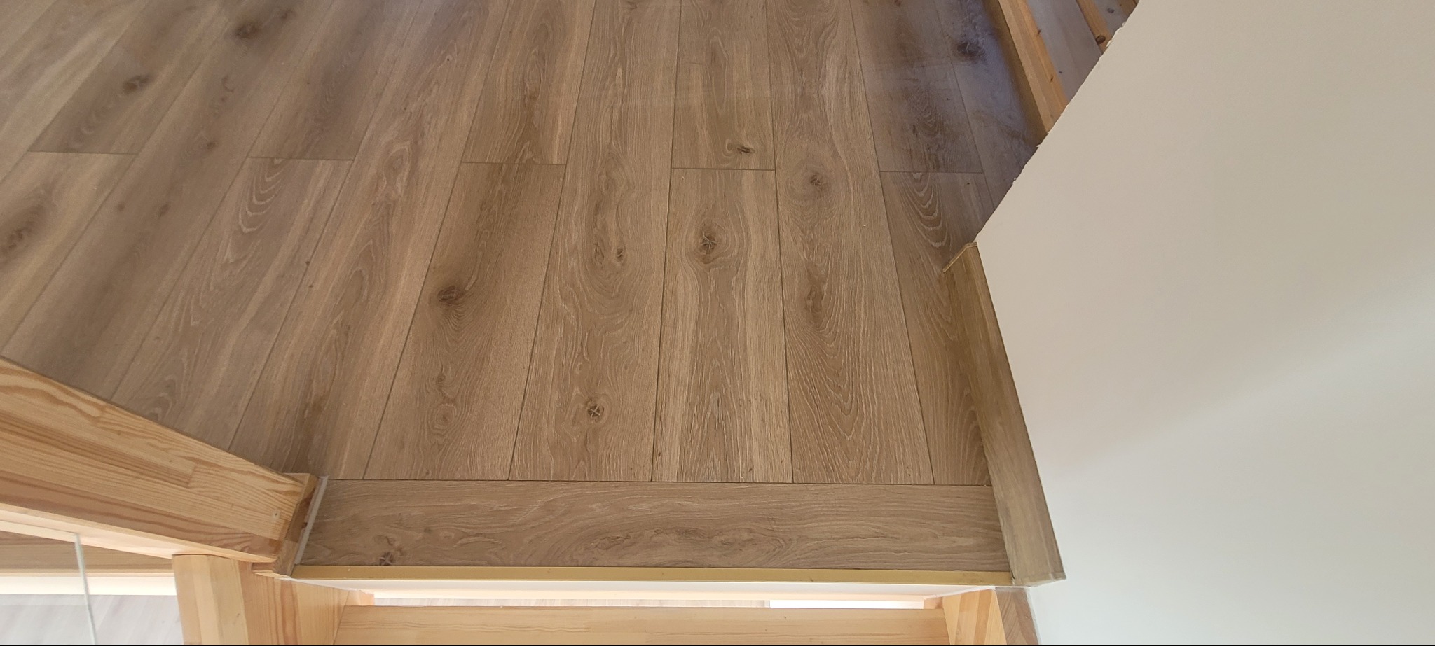 Ułożona podłoga z paneli o wzorze drewna, widok z góry, przy białej ścianie i drewnianej konstrukcji. Widoczne detale łączeń i wykończeń.
