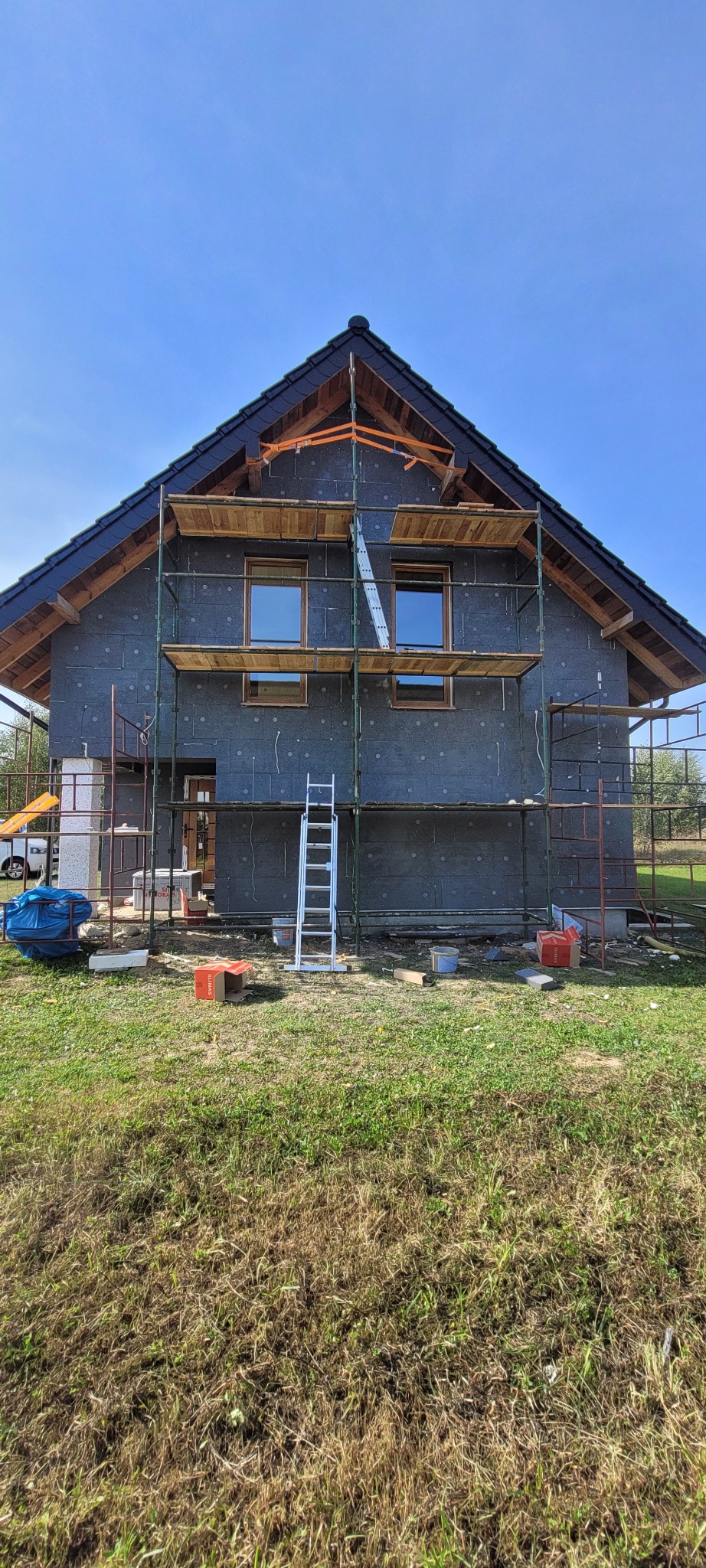 Dom w trakcie ocieplania: szare płyty izolacyjne, rusztowania, drabina. Widoczne okna i fragment dachu. Prace remontowe na zewnątrz budynku.