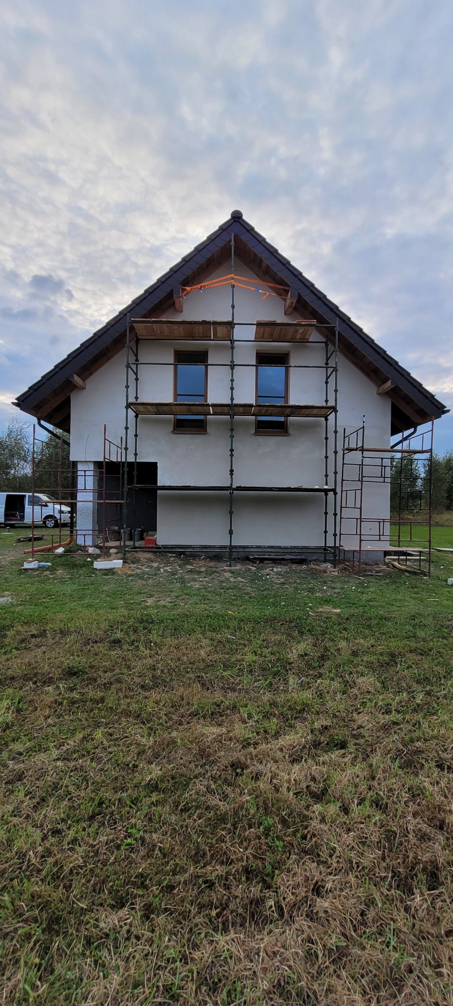 Dom jednorodzinny w trakcie prac wykończeniowych elewacji, z widocznym rusztowaniem. Ściany pokryte świeżym tynkiem, okna w drewnianej ramie.