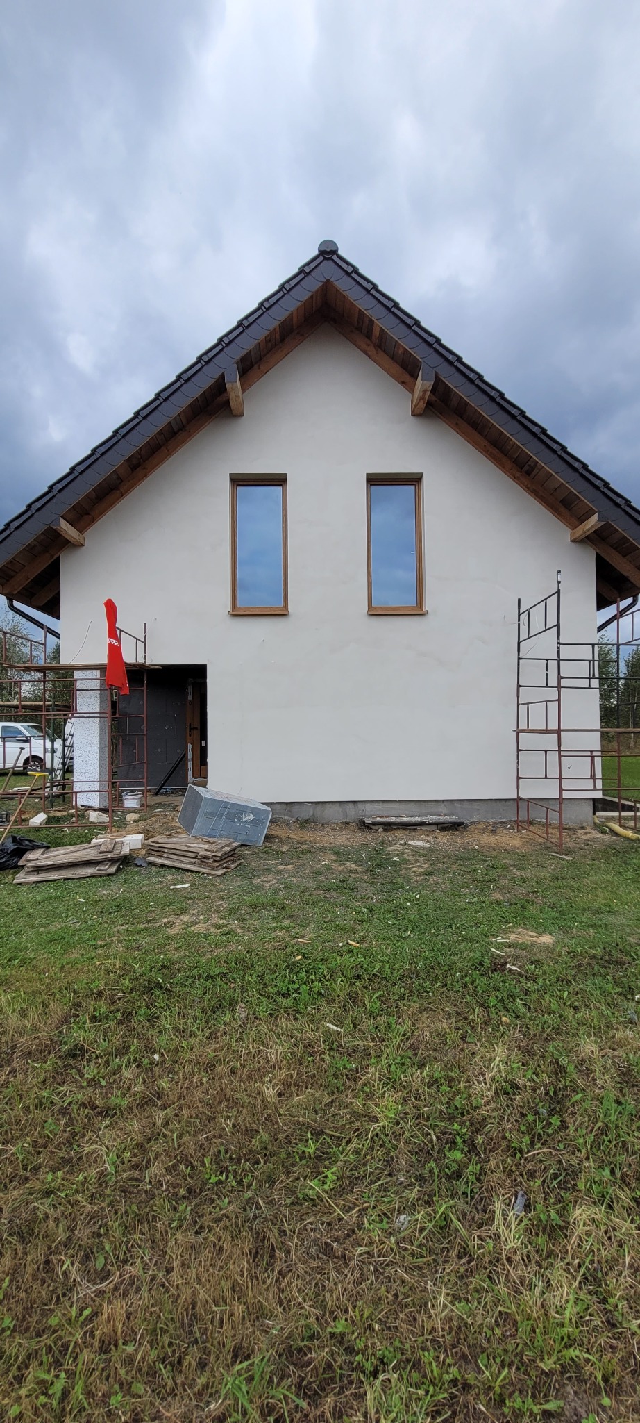Dom jednorodzinny w trakcie prac wykończeniowych. Widoczne rusztowania, świeżo otynkowana elewacja i materiały budowlane na trawniku.