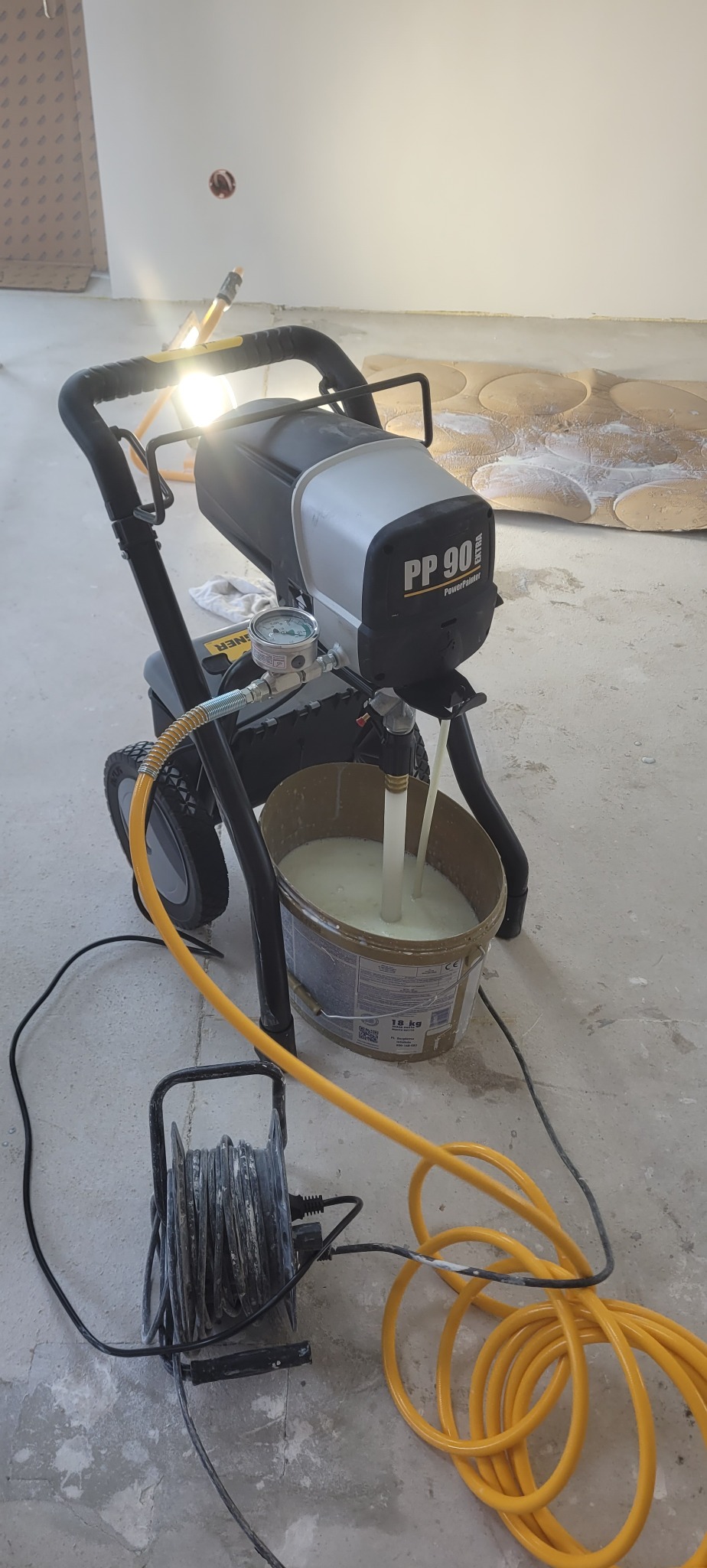 Agregat malarski PP 90 Extra z manometrem, w wiadrze biała masa, obok zwinięty kabel i żółty wąż. Na podłodze folia malarska z wyciętymi otworami.