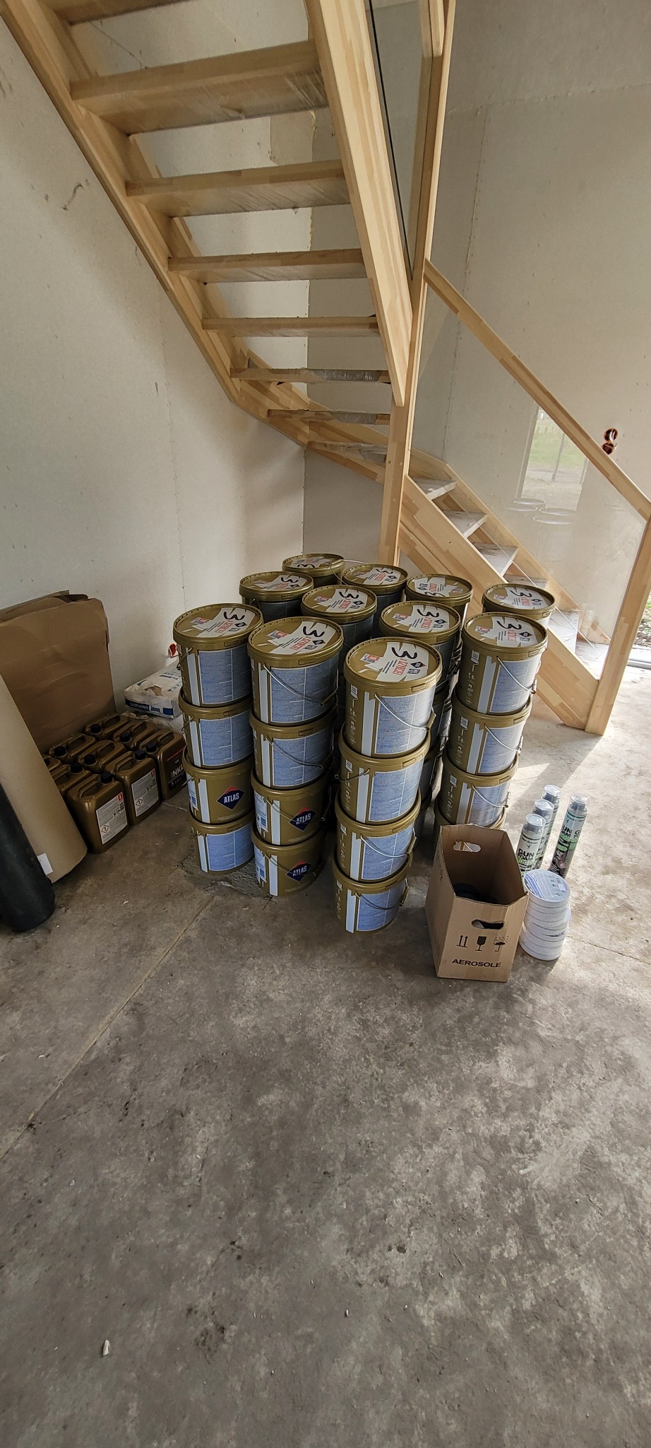 Stos farb Atlas i materiały budowlane pod drewnianymi schodami w surowym wnętrzu. Widok na zaplecze prac wykończeniowych.