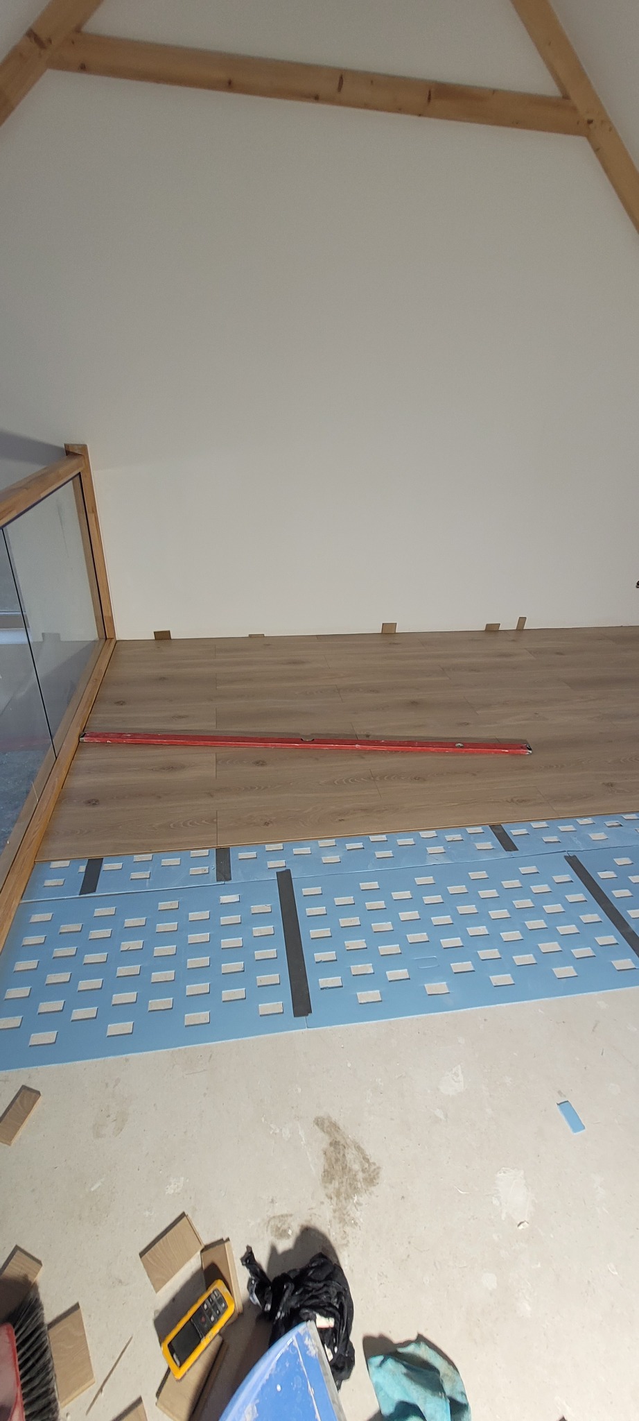 Montaż paneli podłogowych na poddaszu: widoczna izolacja akustyczna i poziomica, częściowo ułożone panele. Elementy wykończeniowe pod ścianą ze szklaną balustradą.