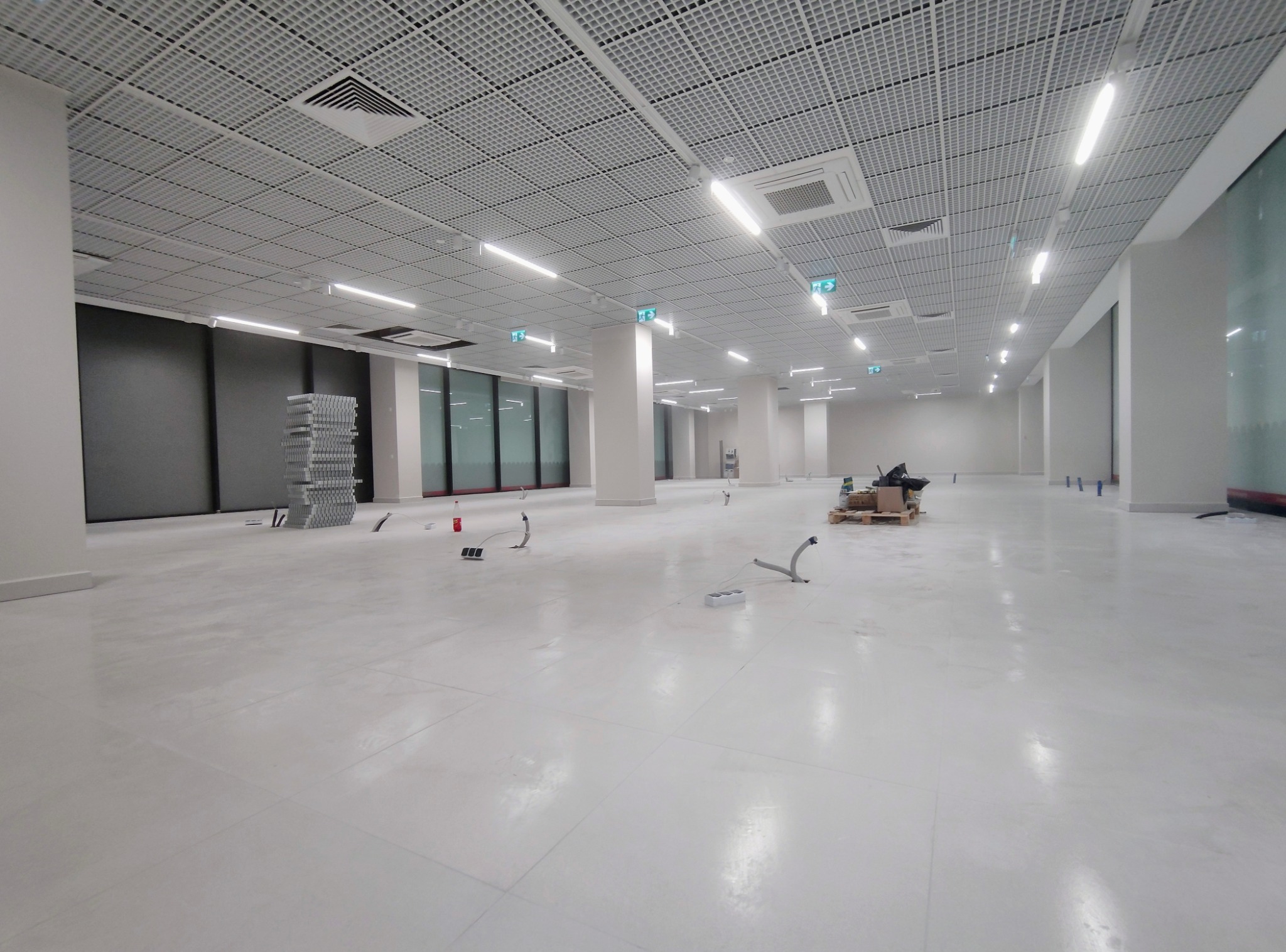 Wnętrze biurowe w trakcie montażu oświetlenia: widoczny sufit podwieszany z rastrem, lampy liniowe, kable i gniazda podłogowe. Stan surowy, przygotowanie do prac wykończeniowych.