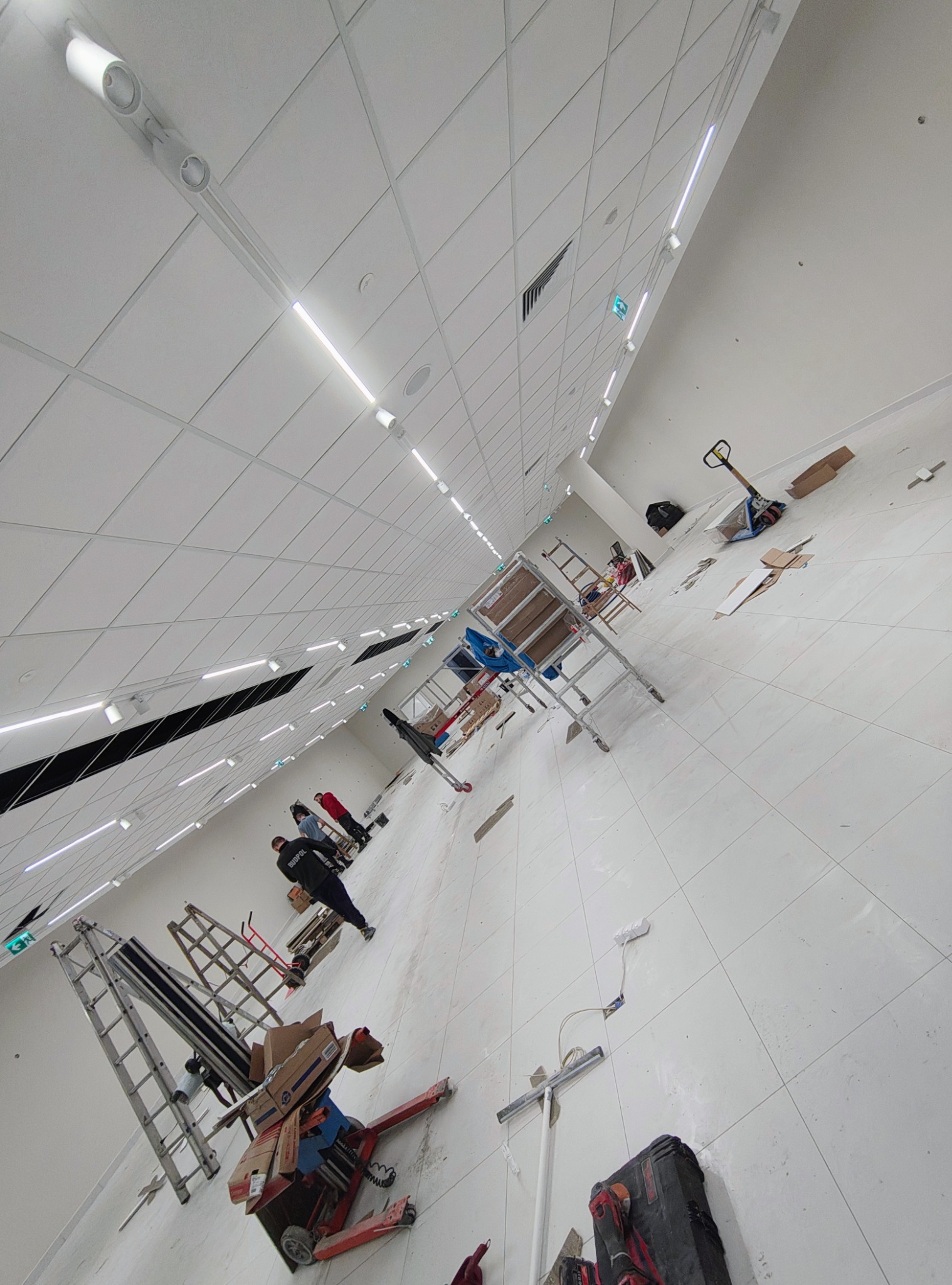 Montaż oświetlenia w dużej hali z białym sufitem kasetonowym. Widoczne lampy liniowe LED, drabiny, narzędzia i osoby w oddali. Ukośny kadr.