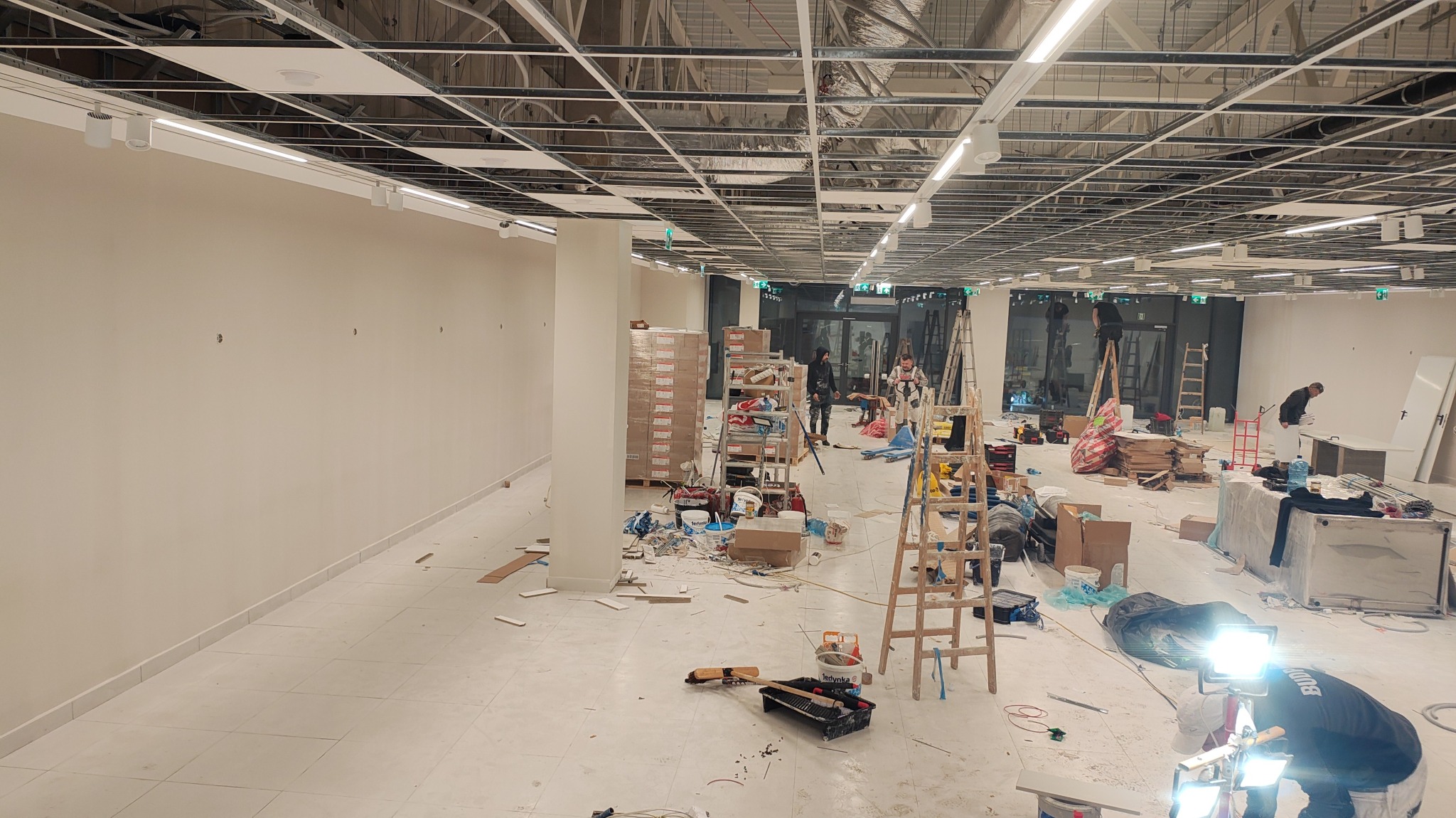 Montaż oświetlenia w przestrzeni komercyjnej: widoczne drabiny, narzędzia i ekipa montażowa. Sufit z widoczną instalacją i zamontowanymi lampami.