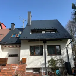 Dom z ciemnoszarym dachem z blachy trapezowej, widoczne okna dachowe i komin. Biała elewacja budynku, brązowe ramy okienne, ogr&oacute;d z przodu.