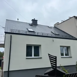 Dom po renowacji z ciemnoszarą, metalową dachówką, oknami dachowymi i świeżo pomalowaną, jasną elewacją. Widoczne detale wykończenia i czarny komin.