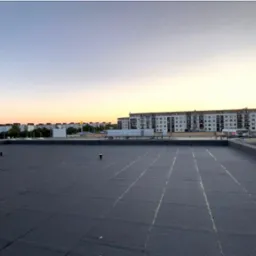 Wykończony płaski dach pokryty czarną papą termozgrzewalną z widocznymi liniami łączeń, na tle zabudowy miejskiej i zachodzącego słońca.