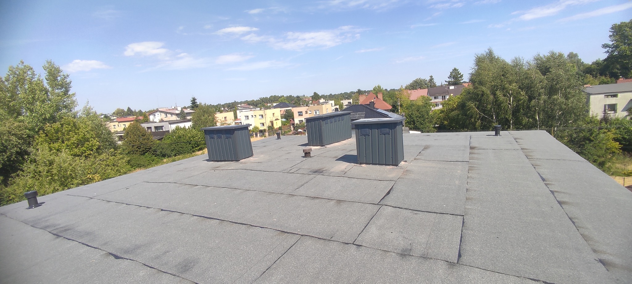 Dach pokryty papą termozgrzewalną w Częstochowie. Widoczne kominki wentylacyjne i zabudowa kominów. Tło: zabudowa miejska i drzewa w słoneczny dzień.