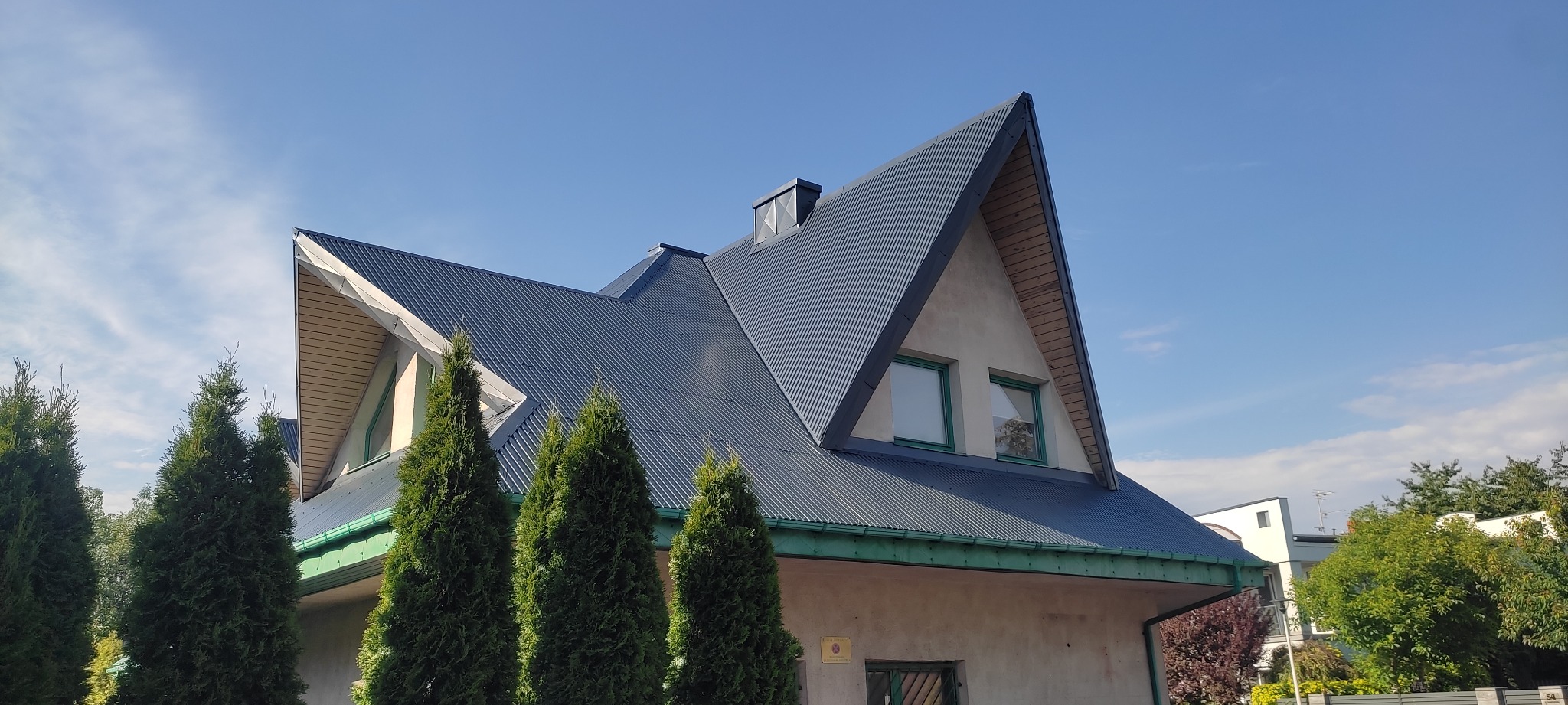 Dom z ciemnym, metalowym dachem po malowaniu, z widocznymi wiatrownicami i rynnami w kolorze zielonym, na tle błękitnego nieba.