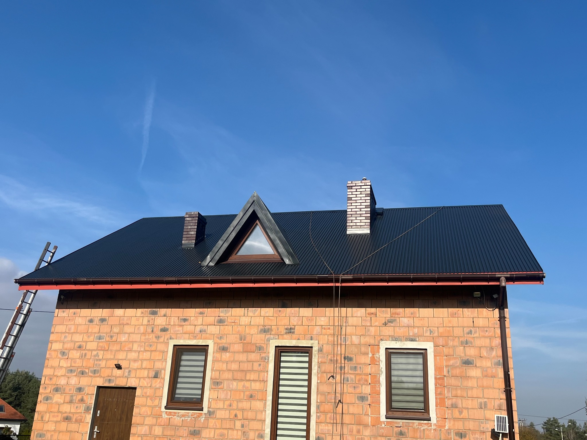 Dom z czarnym dachem z blachy trapezowej, ceglaną elewacją i kominem. Na dachu widoczna drabina. Błękitne niebo w tle.