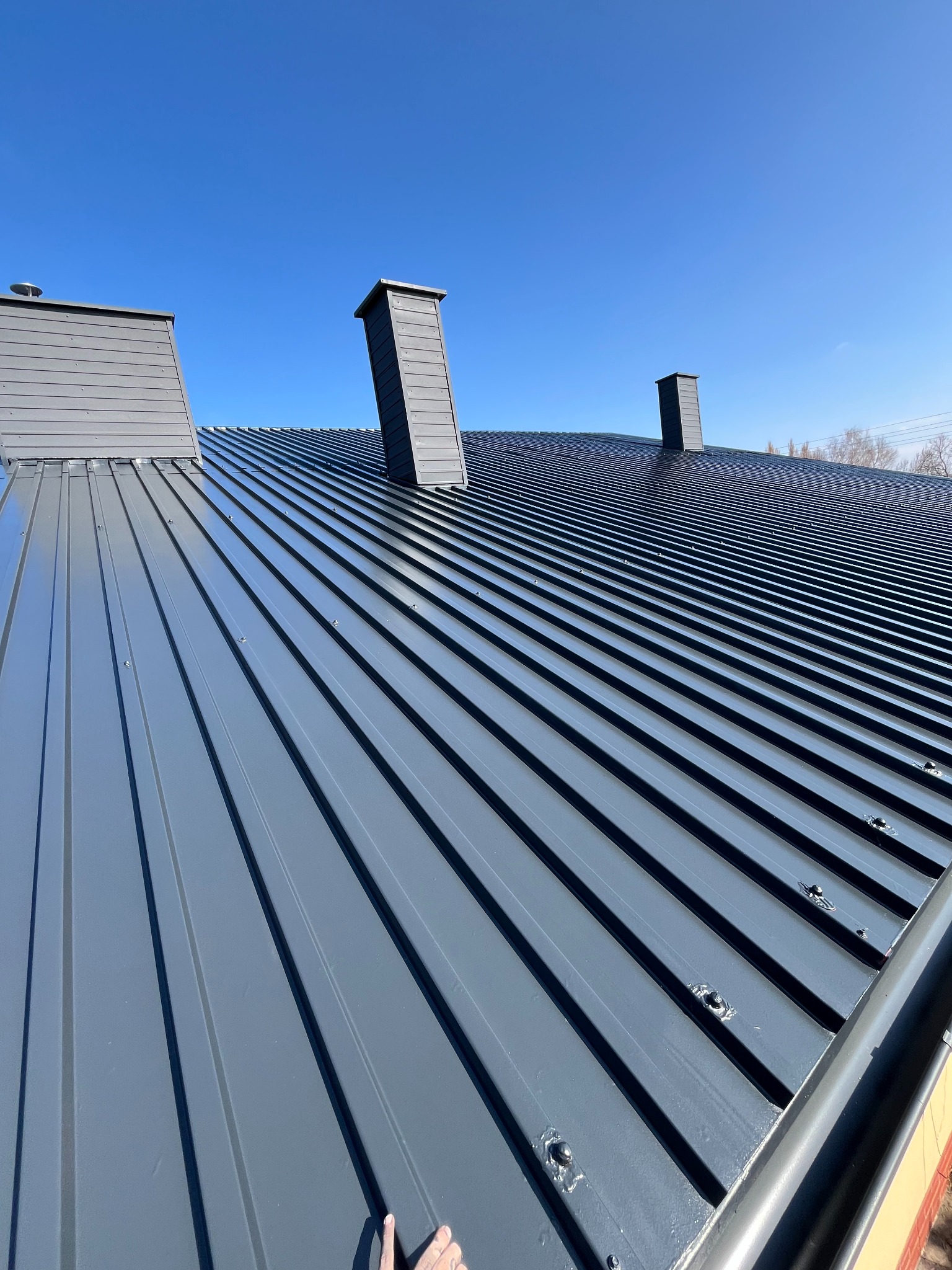 Szary dach z blachy trapezowej, widok z dołu na trzy kominy. Nowe pokrycie dachu z rynnami w słoneczny dzień. Detal łączenia blach wzdłuż połaci dachowej.