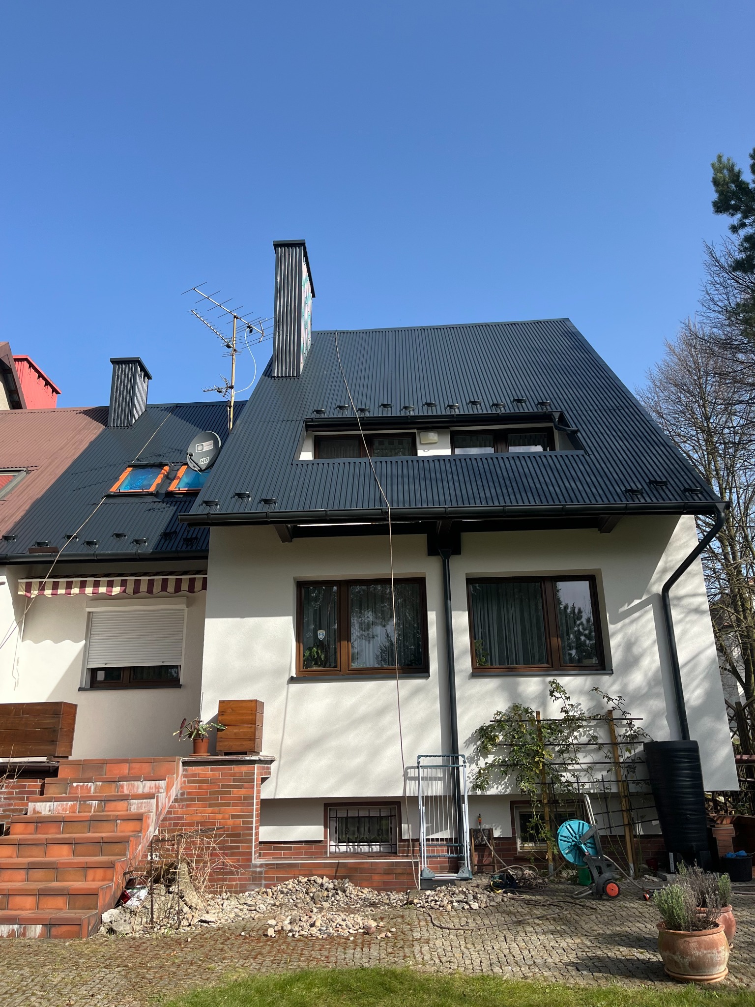 Dom z ciemnoszarym dachem z blachy trapezowej, widoczne okna dachowe i komin. Biała elewacja budynku, brązowe ramy okienne, ogród z przodu.