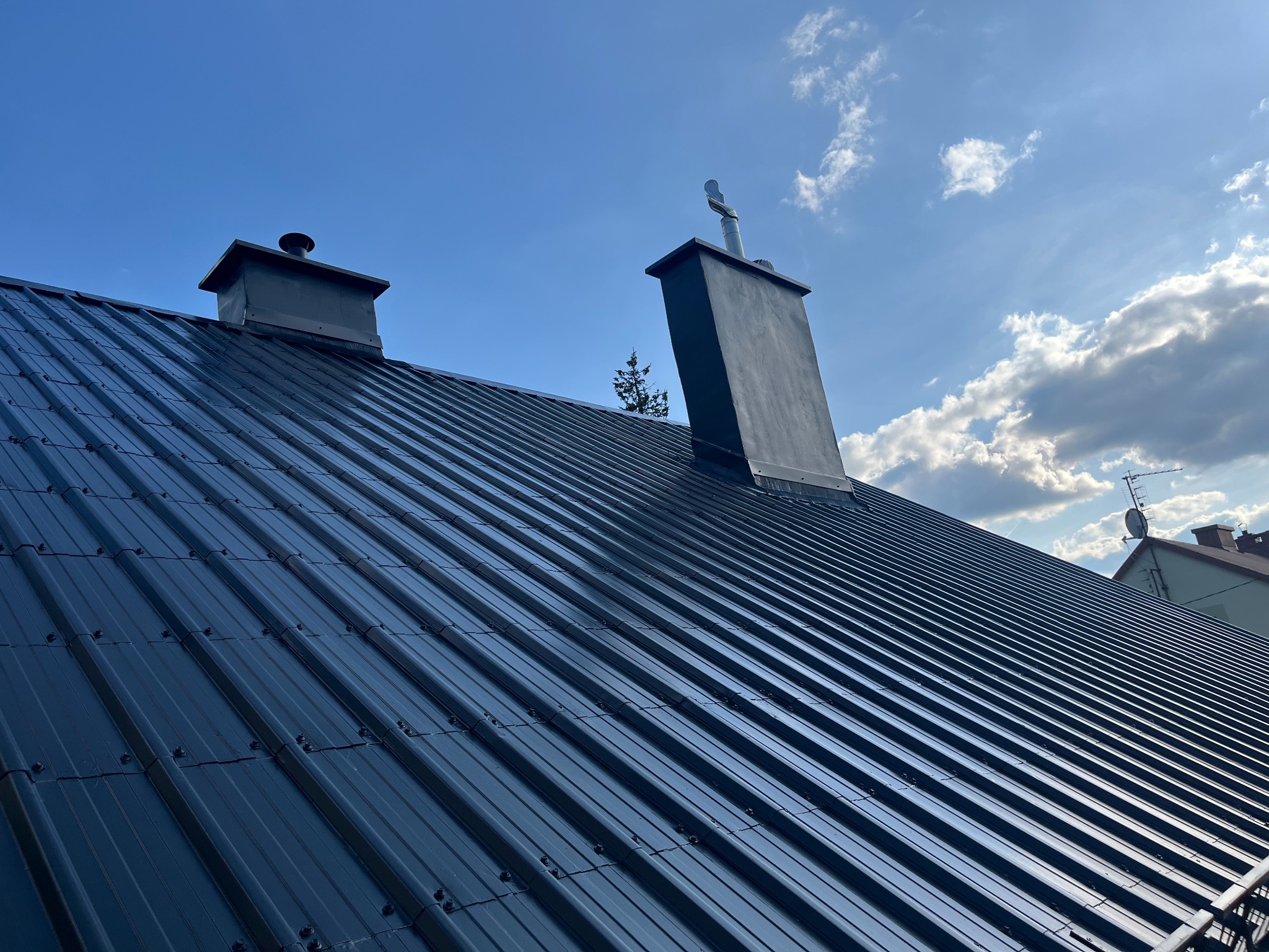 Błyszczący, pomalowany na ciemno dach z blachy trapezowej, z dwoma kominami. Na jednym kominie widoczna metalowa nasada. Błękitne niebo z chmurami w tle.