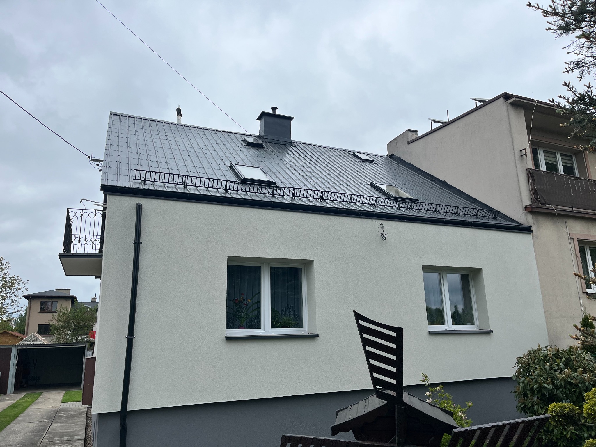Dom po renowacji z ciemnoszarą, metalową dachówką, oknami dachowymi i świeżo pomalowaną, jasną elewacją. Widoczne detale wykończenia i czarny komin.