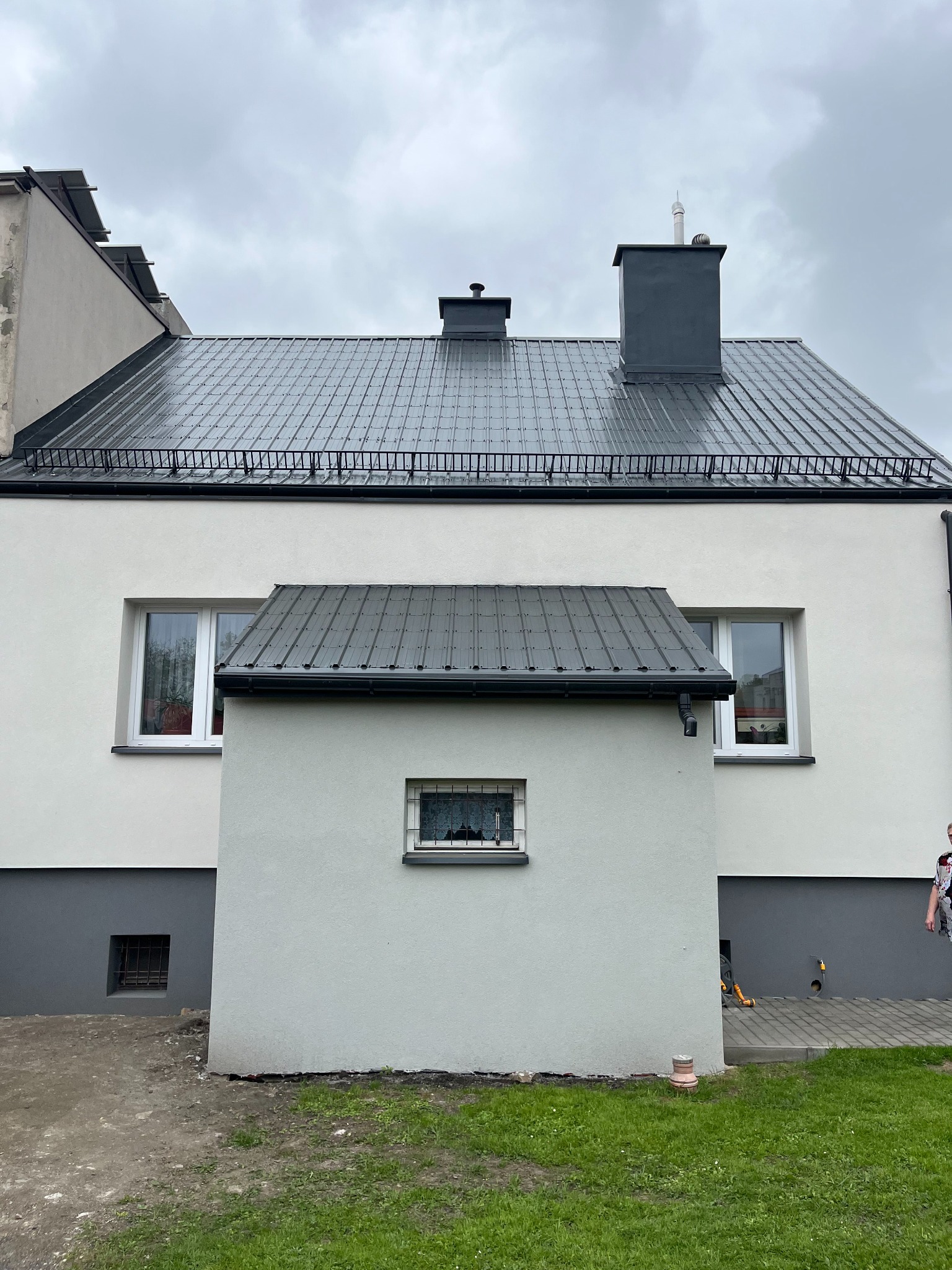 Dom po malowaniu elewacji i dachu na szaro. Widoczny komin, okna i fragment trawnika. Ujęcie z dołu, pochmurne niebo w tle.