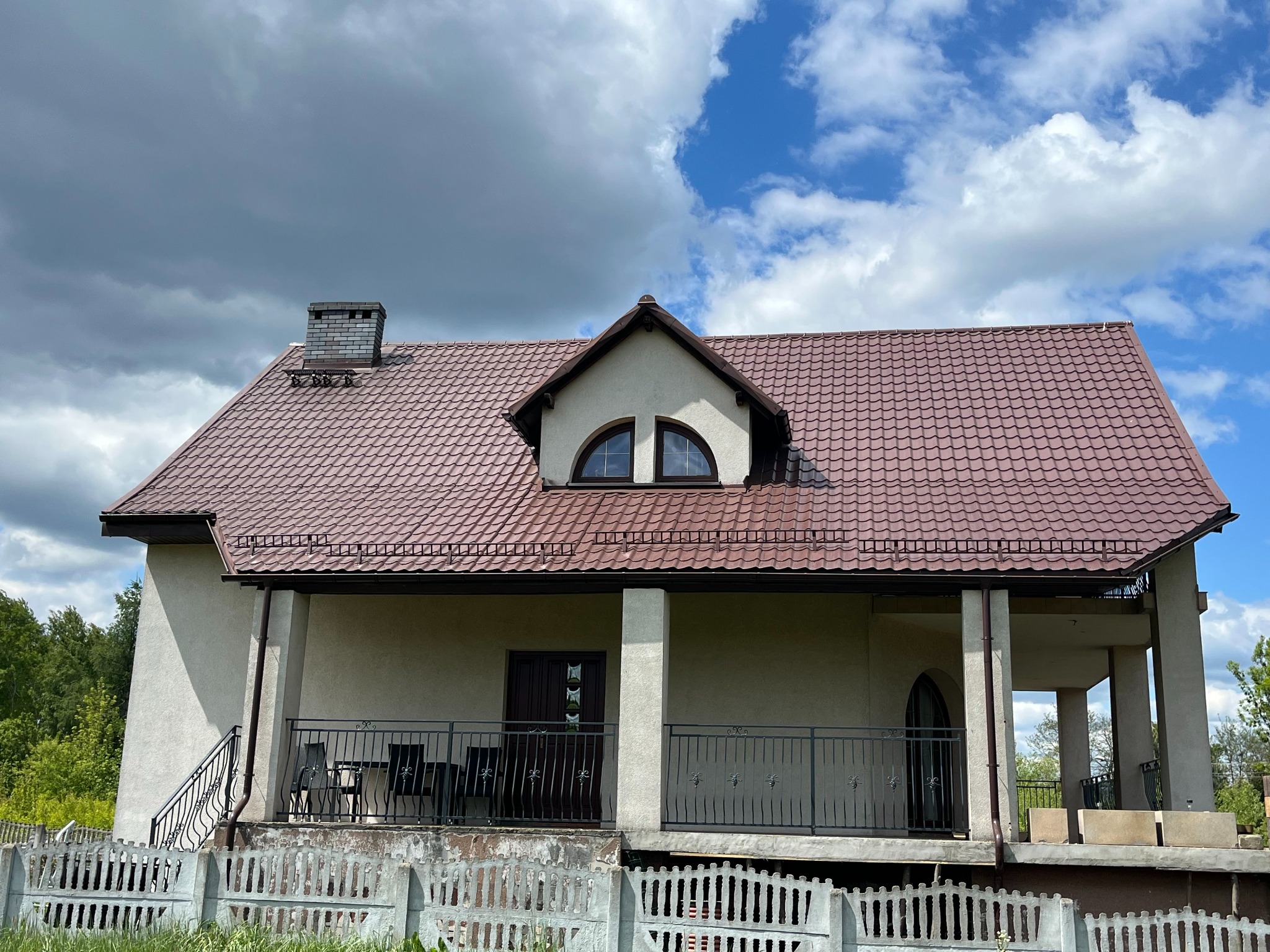 Dom z brązowym dachem, widoczny komin z klinkieru i ława kominiarska. Na pierwszym planie ogrodzenie z betonowych elementów. Błękitne niebo z chmurami w tle.